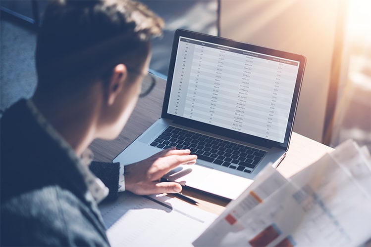 Mann hält Listen in der rechten Hand und arbeitet am Laptop mit Tabellen zur Validierung von Handelsdaten in Power BI.