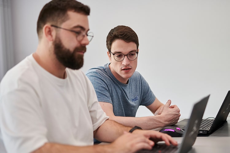 Zwei Männer an einem Schreibtisch mit Laptops, der hintere beugt sich nach vorne und spricht zum anderen – Zusammenarbeit zur Entwicklung eines Fachkonzepts für ein Maklerportal.