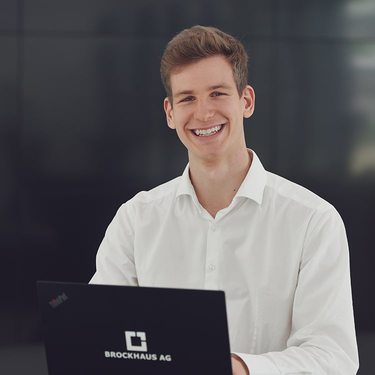 junger BROCKHAUS AG Mitarbeiter lächelt in die Kamera, vor ihm der Laptop
