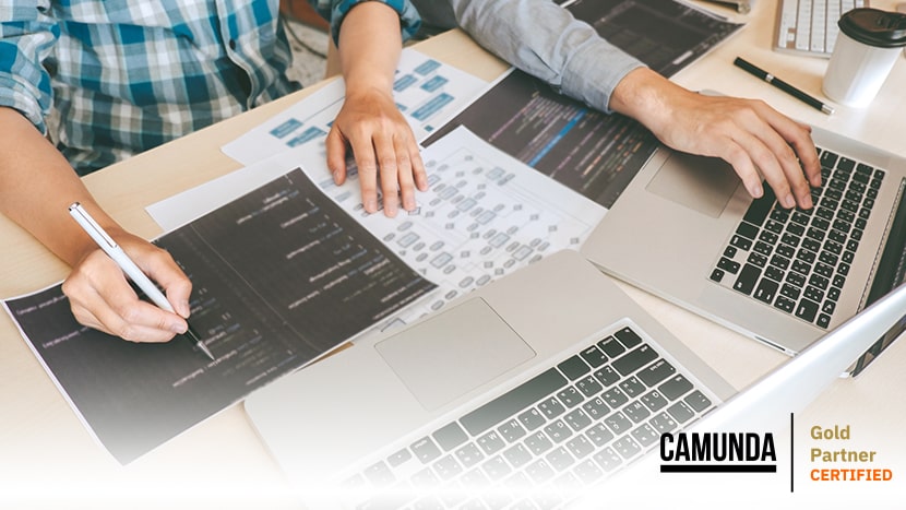Zwei Personen arbeiten an Laptops und analysieren Prozessdiagramme und Softwarecodes auf Papier, um mit Camunda BPM maßgeschneiderte Prozessautomatisierungslösungen zu entwickeln.