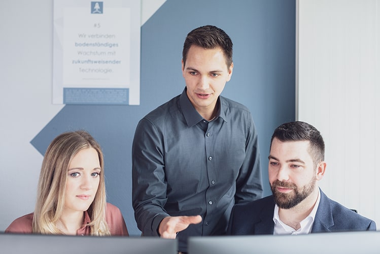 Zwei Mitarbeitende sitzen an einem Schreibtisch und schauen in den Bildschirm, während ein Kollege hinter ihnen steht und auf etwas auf dem Bildschirm zeigt. Sie arbeiten gemeinsam an einem Projekt und besprechen ihre Fortschritte.