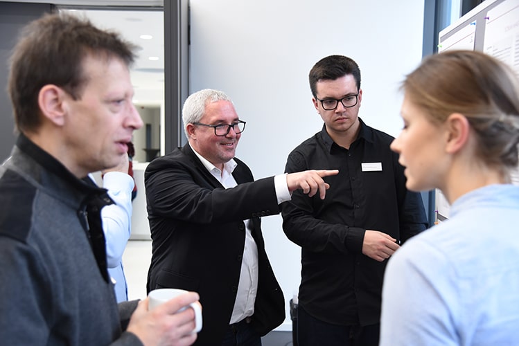 Eine Gruppe von BROCKHAUS Mitarbeitenden steht zusammen. Die zwei Personen im Vordergrund sind ins Gespräch vertieft. Im Hintergrund zeigt ein Mitarbeiter auf eine Metaplanwand.