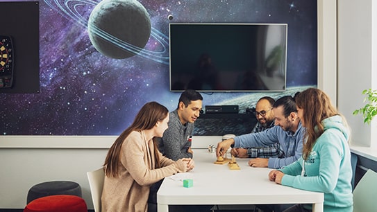 Fünf Mitarbeiter sitzen an einem Tisch und spielen zusammen ein Spiel. Im Hintergrund sieht man eine Tapete mit Planeten und einen Fernseher an der Wand
